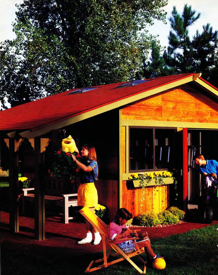 Front corner view of the yard shed built, with plants, porch, and life surrounding it